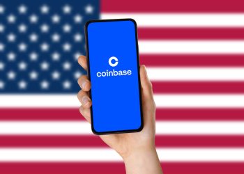 A hand holding a smartphone and the coinbase app logo. The USA flag in the background. coinbase Application Logo. Technology concept. Close up.