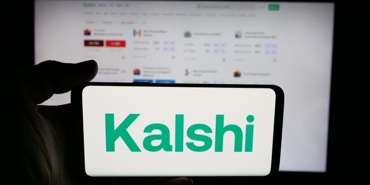 Person holding cellphone with logo of US prediction market company Kalshi Inc. on screen in front of webpage. Focus on phone display.